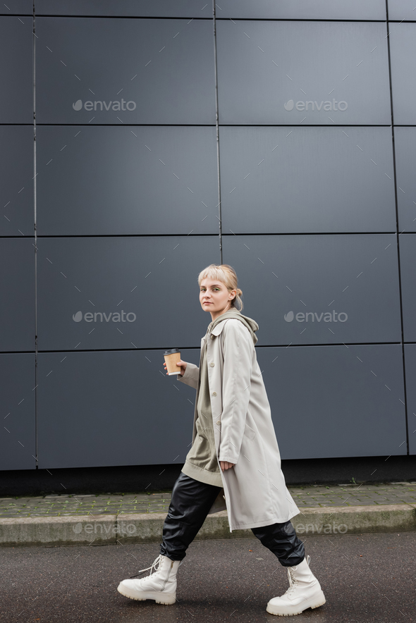 full length of trendy woman with blonde hair with bangs walking in coat, black leather pants