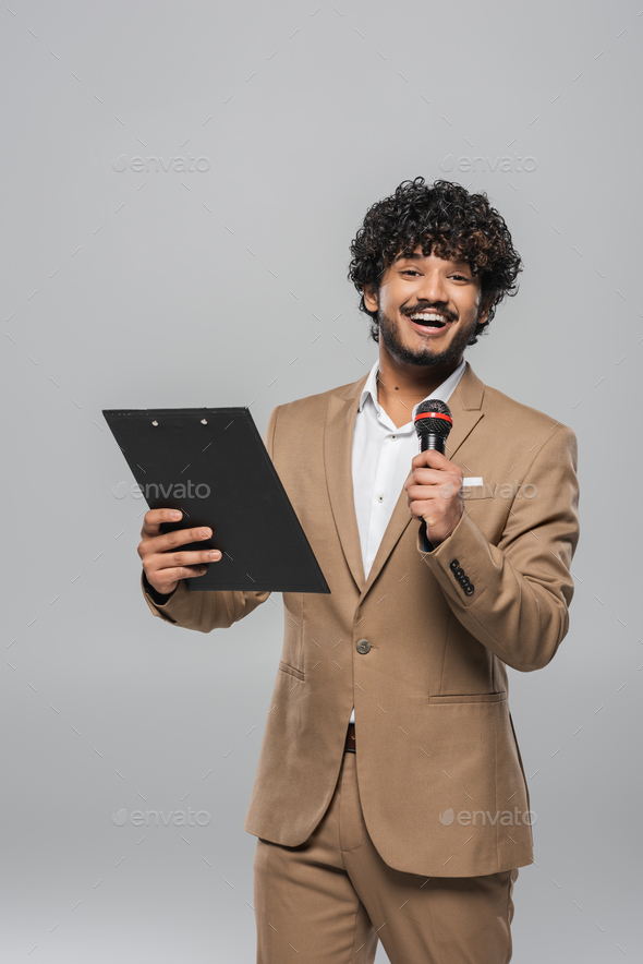 Cheerful and curly young indian host of event holding microphone and clipboard while looking