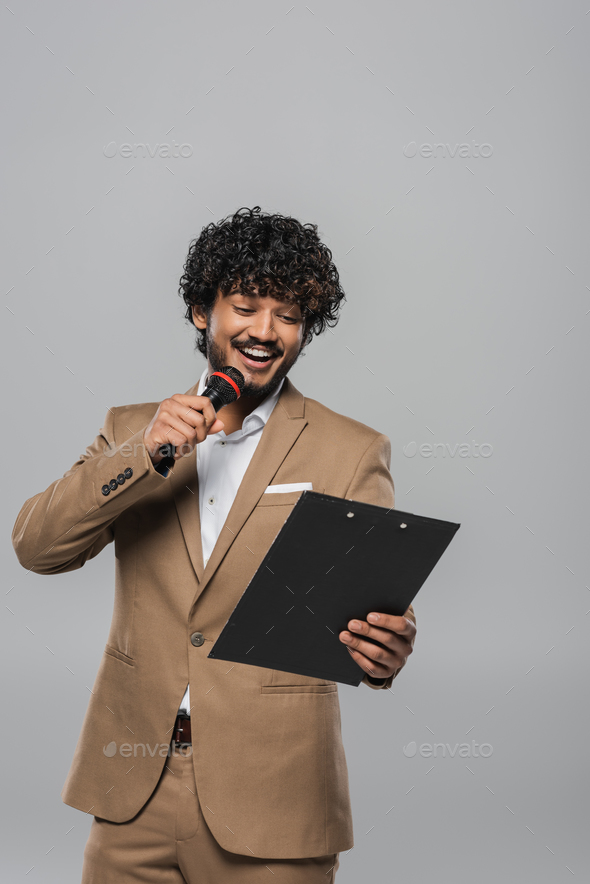 Young indian host of event in formal wear smiling while talking at microphone and holding clipboard