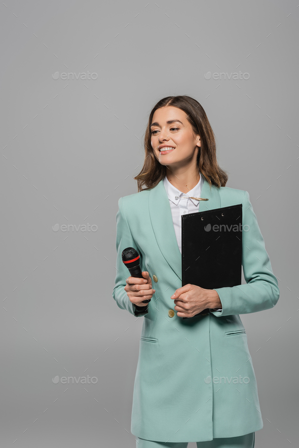 Cheerful brunette host of event in blue jacket and shirt holding microphone and clipboard