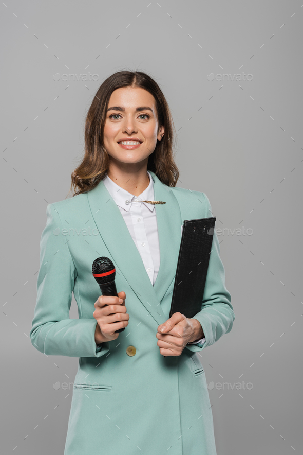 Portrait of positive brunette host of event in blue suit holding microphone and clipboard