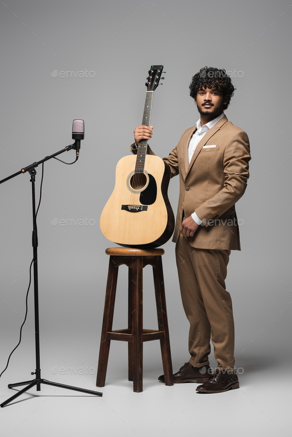 Full length of indian host of event in formal wear holding acoustic guitar near chair