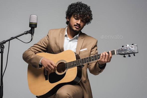 Young and curly indian host of event playing acoustic guitar near microphone on stand while sitting