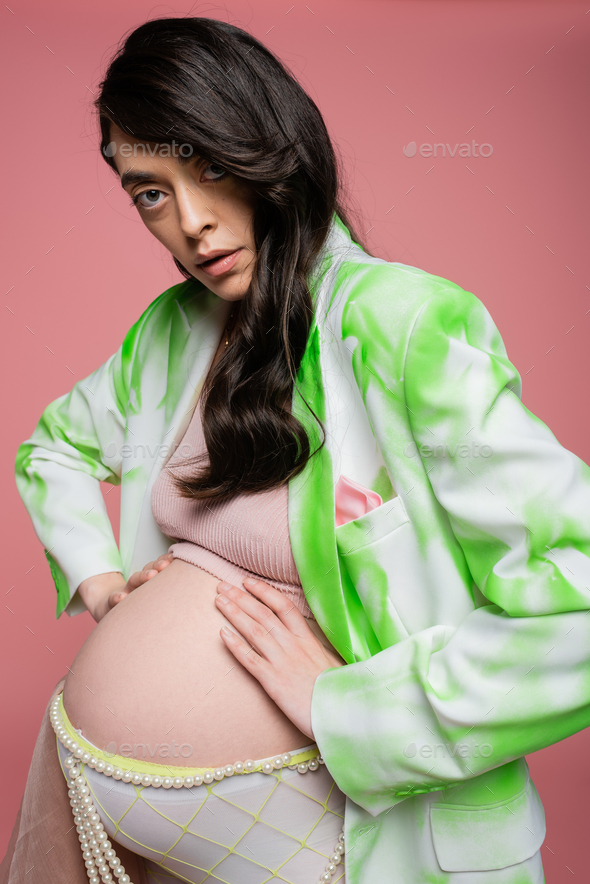 pregnant woman with wavy brunette hair, wearing green and white blazer,  crop top and beads belt Stock Photo by LightFieldStudios