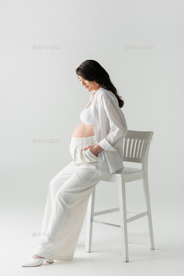 Side view of cropped serene unrecognizable pregnant female in underwear  sitting on stool in room with green houseplants and tenderly touching tummy  stock photo