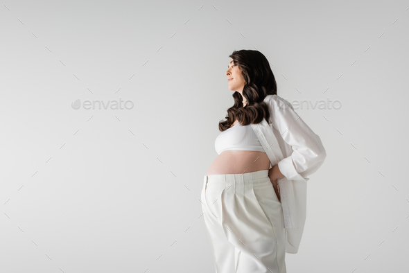 side view of smiling pregnant woman with wavy brunette hair, wearing white stylish maternity