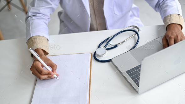 Medical professional in a lab coat holding a pencil on an empty file and operating a laptop.