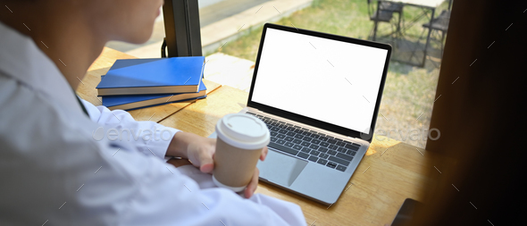 View over shoulder of doctor holding paper cup of coffee and using laptop.