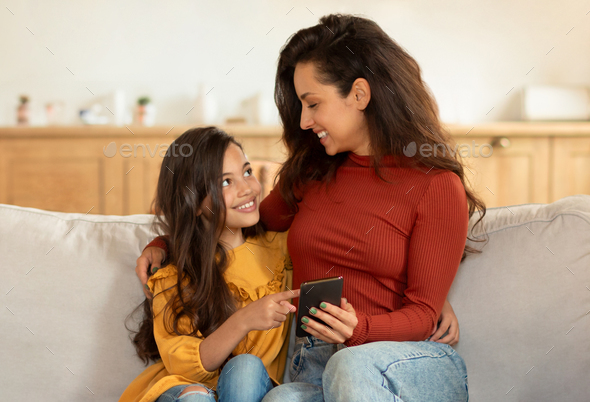 Happy Middle Eastern Mom And Little Daughter Using Smartphone Indoor