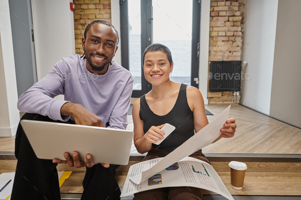 happy man and woman holding gadgets and graphs, interracial colleagues looking at camera, startup