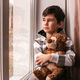 A sad boy looks out the window at the rainy autumn weather, hugging a teddy bear. - PhotoDune Item for Sale