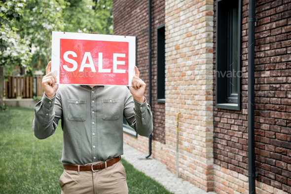 real estate agent obscuring face with sale signboard while standing near cottage in city