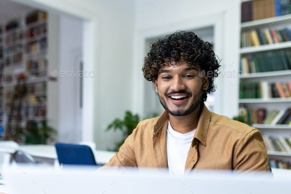 Indian student with curly hair studying sitting among books on shelves, man watching video course