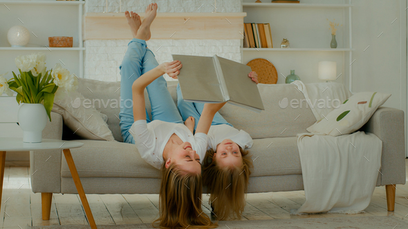 Cheerful Caucasian mother lying upside down on comfortable sofa with little child girl kid daughter