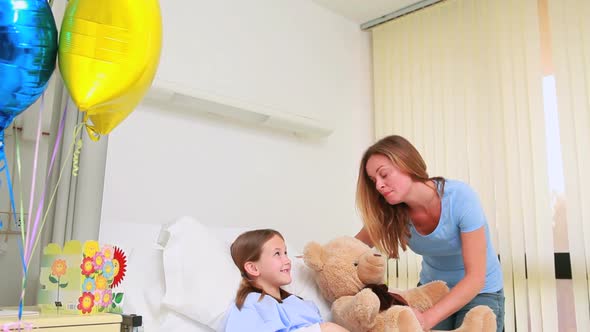 Smiling woman giving a teddy bear to a smiling girl in a bed alt