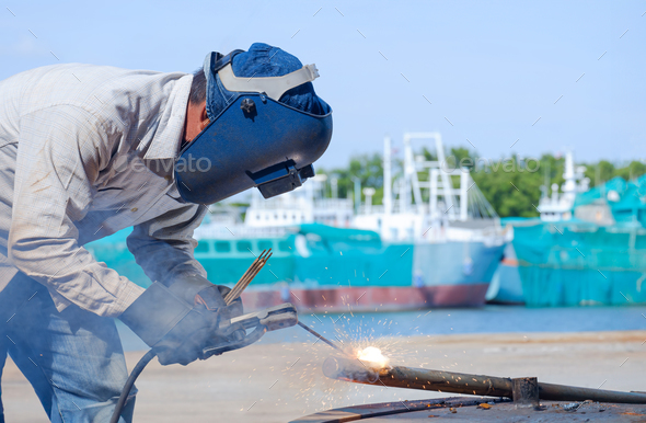 Welder with safety equipment is welding galvanized steel pipe in shipyard area at harbor
