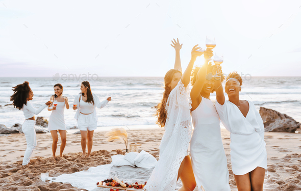 Carefree ladies bride team toasting wine glasses while having hen party and dancing on the beach at