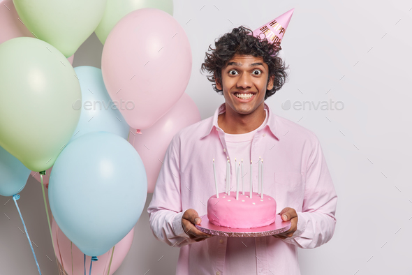Spoiled holiday concept. Upset gloomy man poses dirty with birthday cake  and rosy helium balloons ha Stock Photo by wayhomestudioo