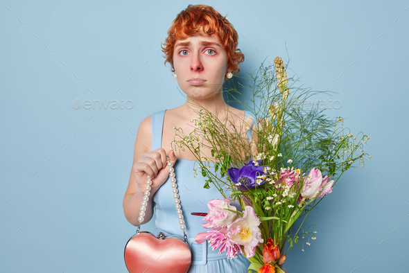 Photo of redhead young European woman has red watery eyes holds small heart shaped bag and bouquet