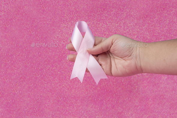 Woman hand holding a light pink ribbon on a pink background - cancer symbol  Stock Photo by wirestock