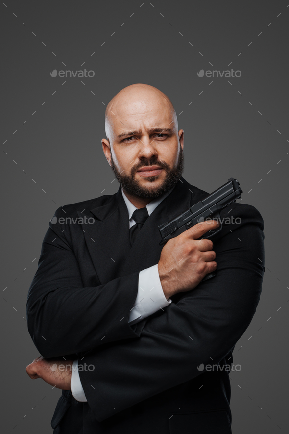 Bald and bearded man in a black suit holds a pistol, exuding power