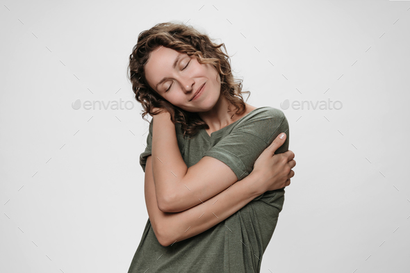 Young curly woman hugging herself, looks happy, loves herself, has high self esteem