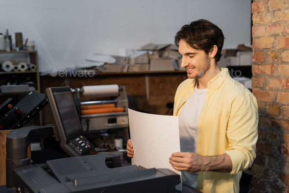 happy publisher holding blank paper near professional equipment in print center