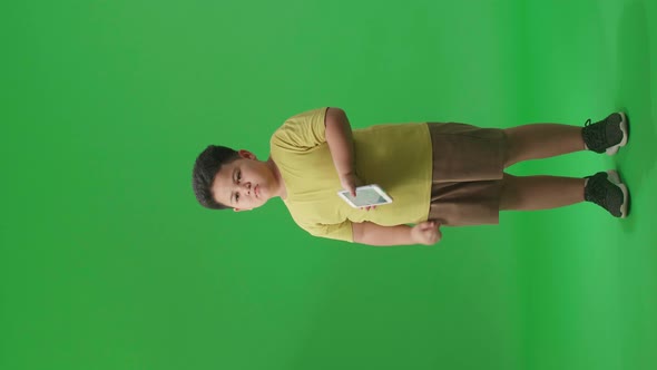 Full Body Of Bored Asian Little Boy Using Mobile Phone In Green Screen Studio alt