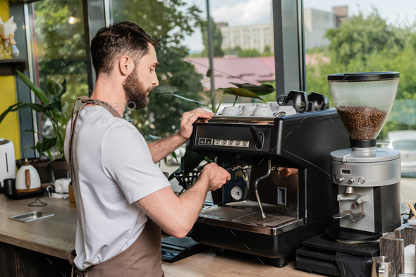 bearded barista in apron using shaker while working near coffee machine in  coffee shop Stock Photo by LightFieldStudios
