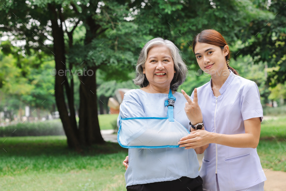 Senior Asian woman wearing patient gown and soft splint due to sore arm showing okay sign with nurse