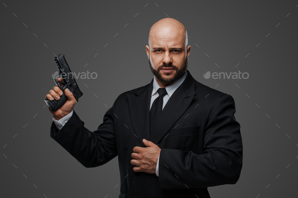 Bald and bearded man in a black suit holds a pistol, exuding power