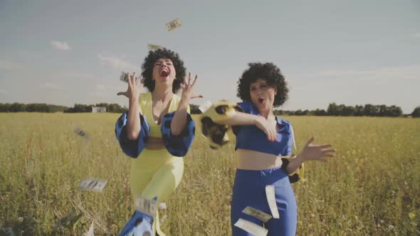 Two Pretty Cheerful Girls in Bright Disco Suits Dance and Have Fun in the Field Under Falling