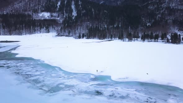 Two people are standing in a big snow field in a valley with a icey lake. alt