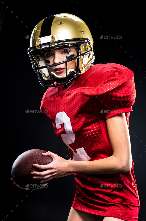 Beautiful female american football player in sportswear and helmet holding  ball on black Stock Photo by LightFieldStudios