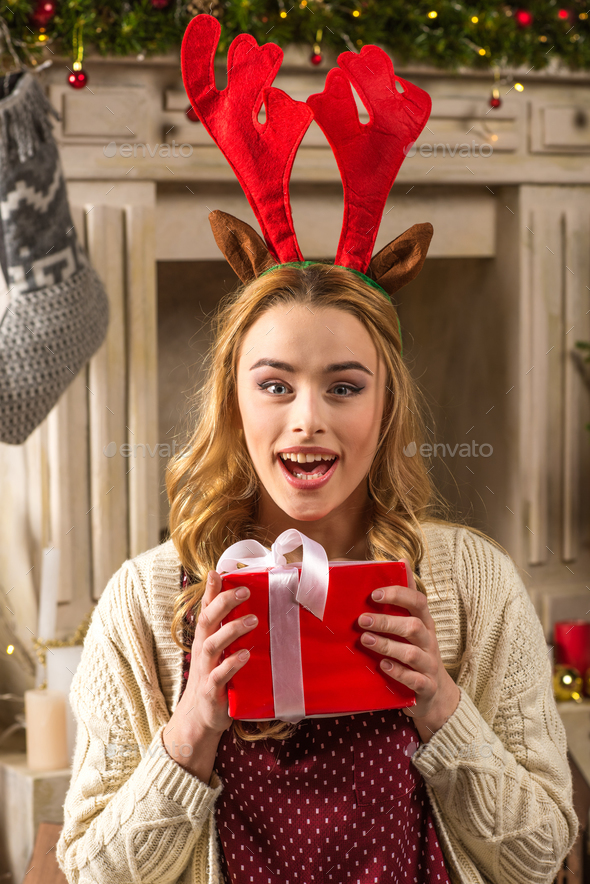 Surprised woman in red antlers hat holding christmas present and looking at camera