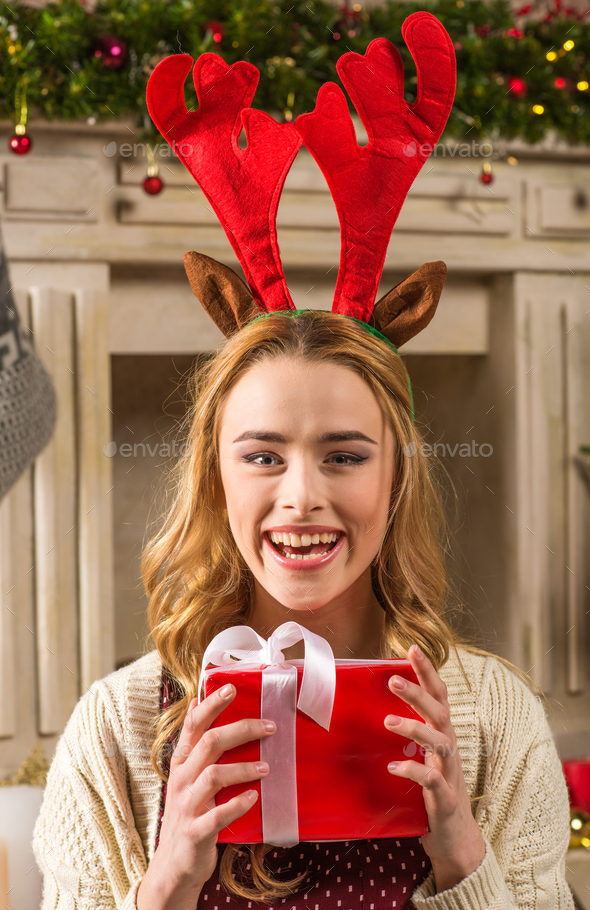 Excited woman in red antlers hat holding christmas present and looking at camera