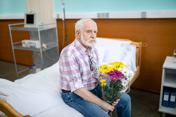 Upset senior man with bouquet sitting on hospital bed and looking away  Stock Photo by LightFieldStudios
