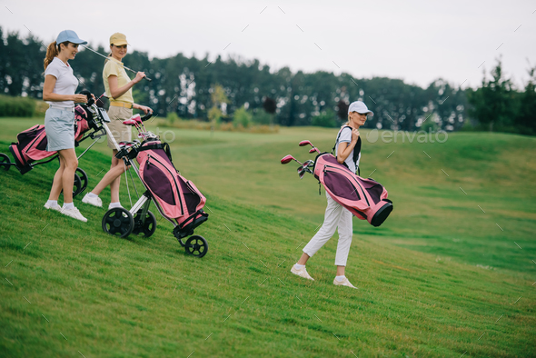 side view of women in polos and caps with golf gear walking on green lawn at golf course