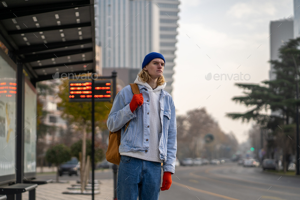 Young man worker waits at bus stop pending for transport coach taxi. Public urban transportation.