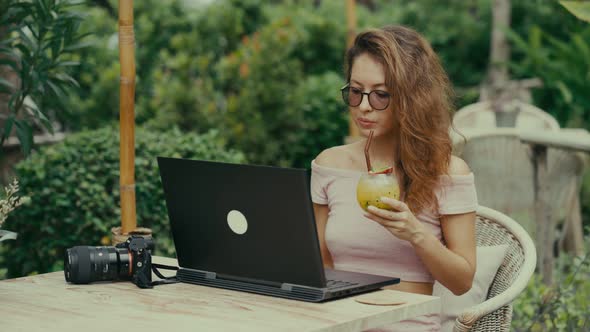 A Young Woman Photographer Drinks a Cocktail While Working with Her Laptop in an Outdoor Cafe alt