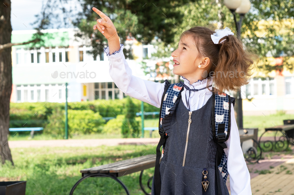 Cheerful funny girl in a school uniform with white bows in school yard points with his finger. Back