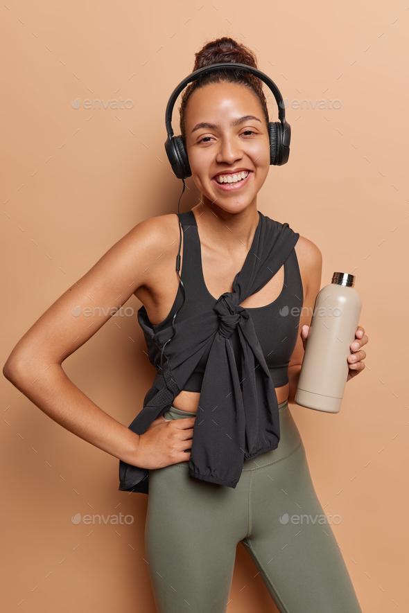 Slim active Latin woman standing with bottle of water at studio after exercising listens music via