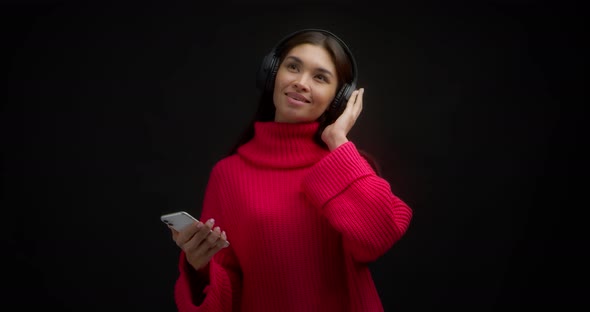 Woman in a Bright Pink Sweater Listens to Music with Headphones Through Phone alt