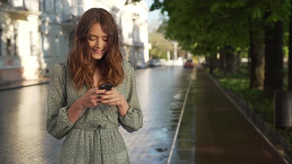 Portrait of Satisfied Young Woman Wearing Dress Smiling and Holding Mobile Phone While Walking alt