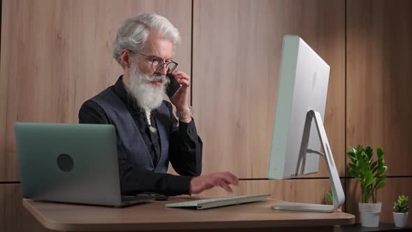 Adult Grayhaired Man Works in the Office at the Computer Typing and Talking on a Mobile Phone an alt
