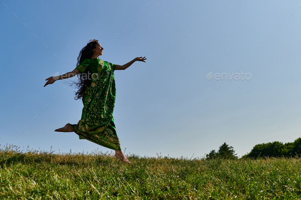 free minded indian woman in sari running on green meadow under blue sky, happy summer