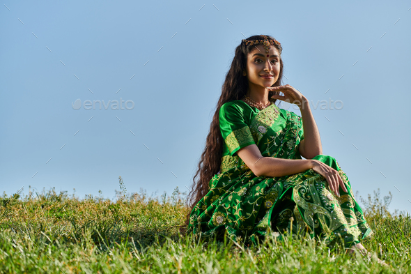 indian woman in ethnic wear, sari, sitting on green lawn under blue summer sky and smiling at camera