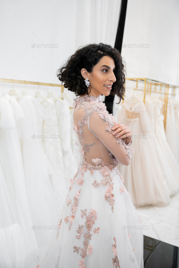cheerful middle eastern and brunette woman with wavy hair standing in gorgeous and floral wedding