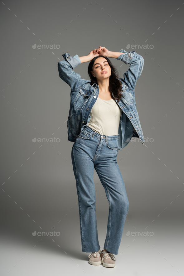 full length of chic woman with brunette hair posing with hands above head and closed eyes