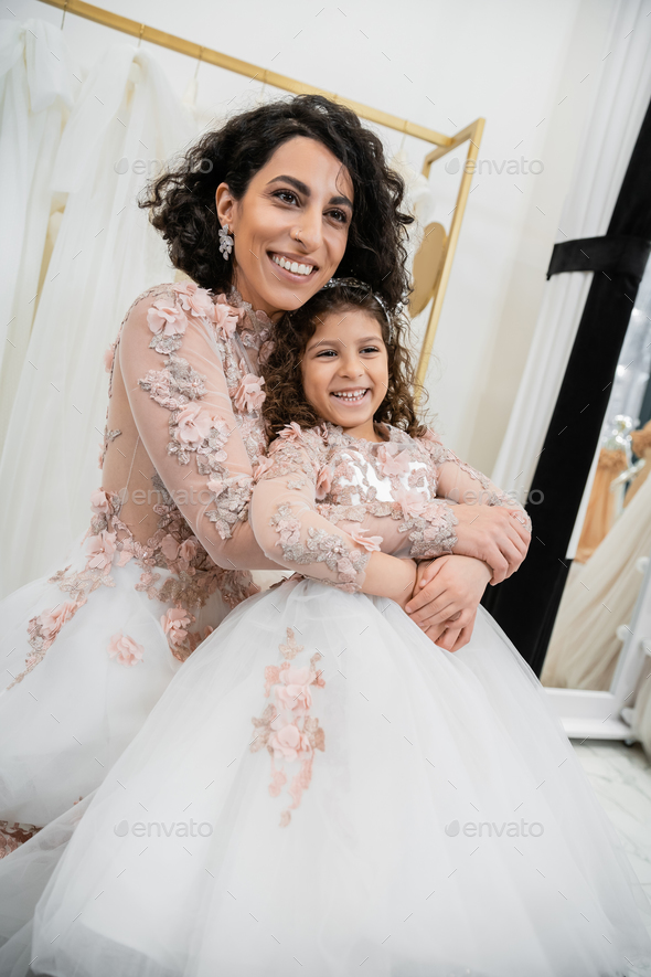 delightful middle eastern bride in wedding dress hugging happy girl in cute  floral attire in bridal Stock Photo by LightFieldStudios